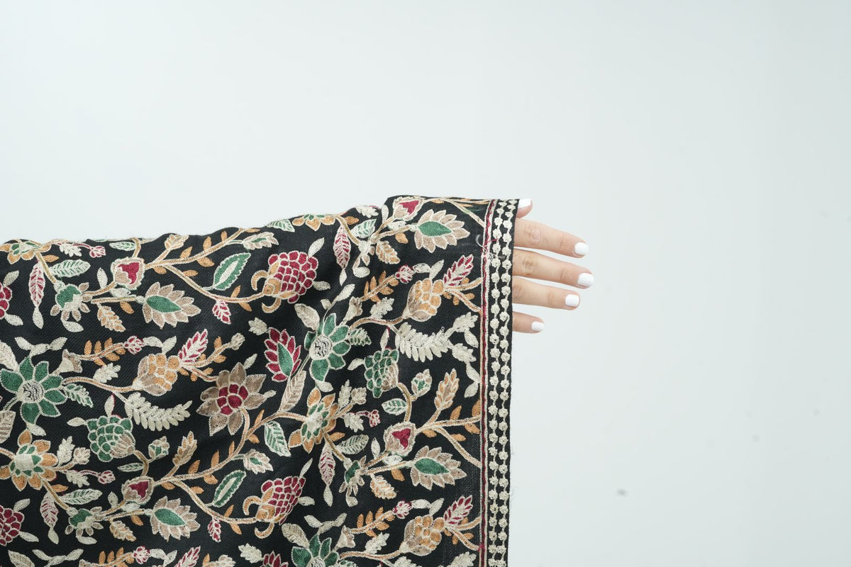 A close-up of a black fabric sleeve or garment featuring an intricate multicolor floral and leaf pattern in beige, green, red, and blue, held out by a hand with white nail polish against a plain white background.
