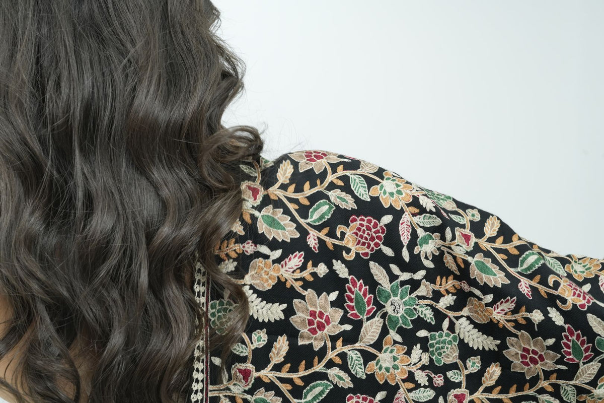 A close-up side view of a person with long, wavy dark brown hair wearing a black embroidered top with intricate floral patterns in red, green, beige, and white.