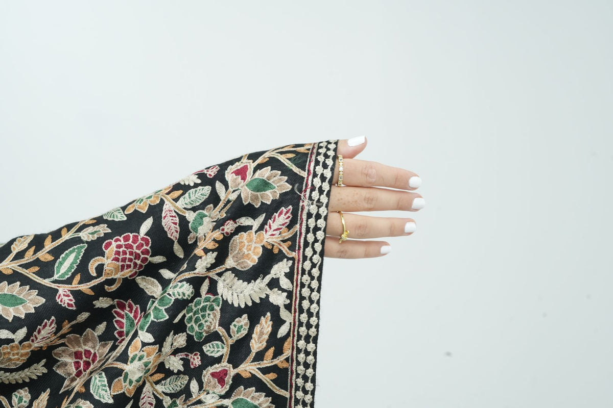 A close-up of a hand with white manicured nails resting on the edge of a black dupatta or scarf featuring intricate multicolor floral and vine embroidery.