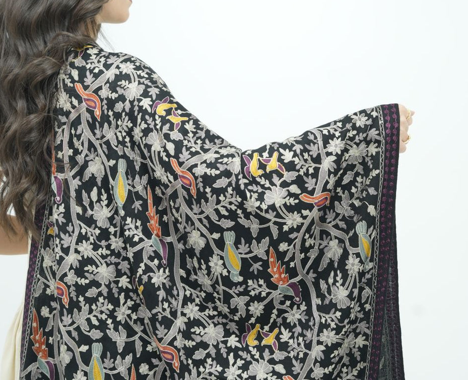 A woman shown from the back wearing a black embroidered shawl with intricate multicolor floral and bird patterns, set against a plain white studio background.