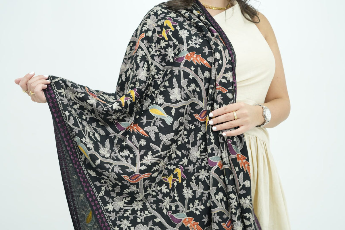 A woman wearing a cream-colored sleeveless dress holds up a black printed shawl or scarf featuring colorful birds and floral motifs. The background is a clean white studio backdrop.