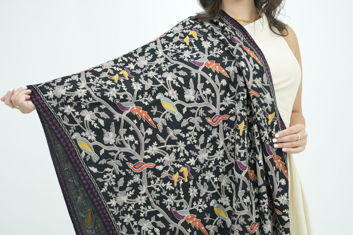 A woman holds up a black printed shawl or scarf featuring a detailed pattern of birds, flowers, and branches in gray, orange, yellow, and purple, set against a plain white background.