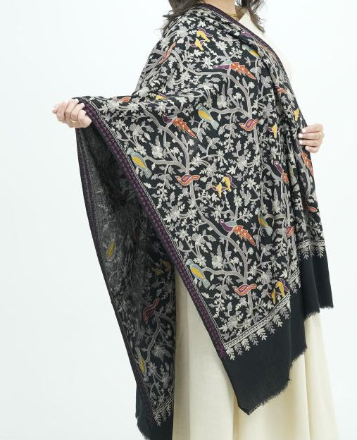 A woman modeling a black shawl with a detailed multicolor floral pattern and a solid black border, worn over a cream-colored dress in a studio setting.