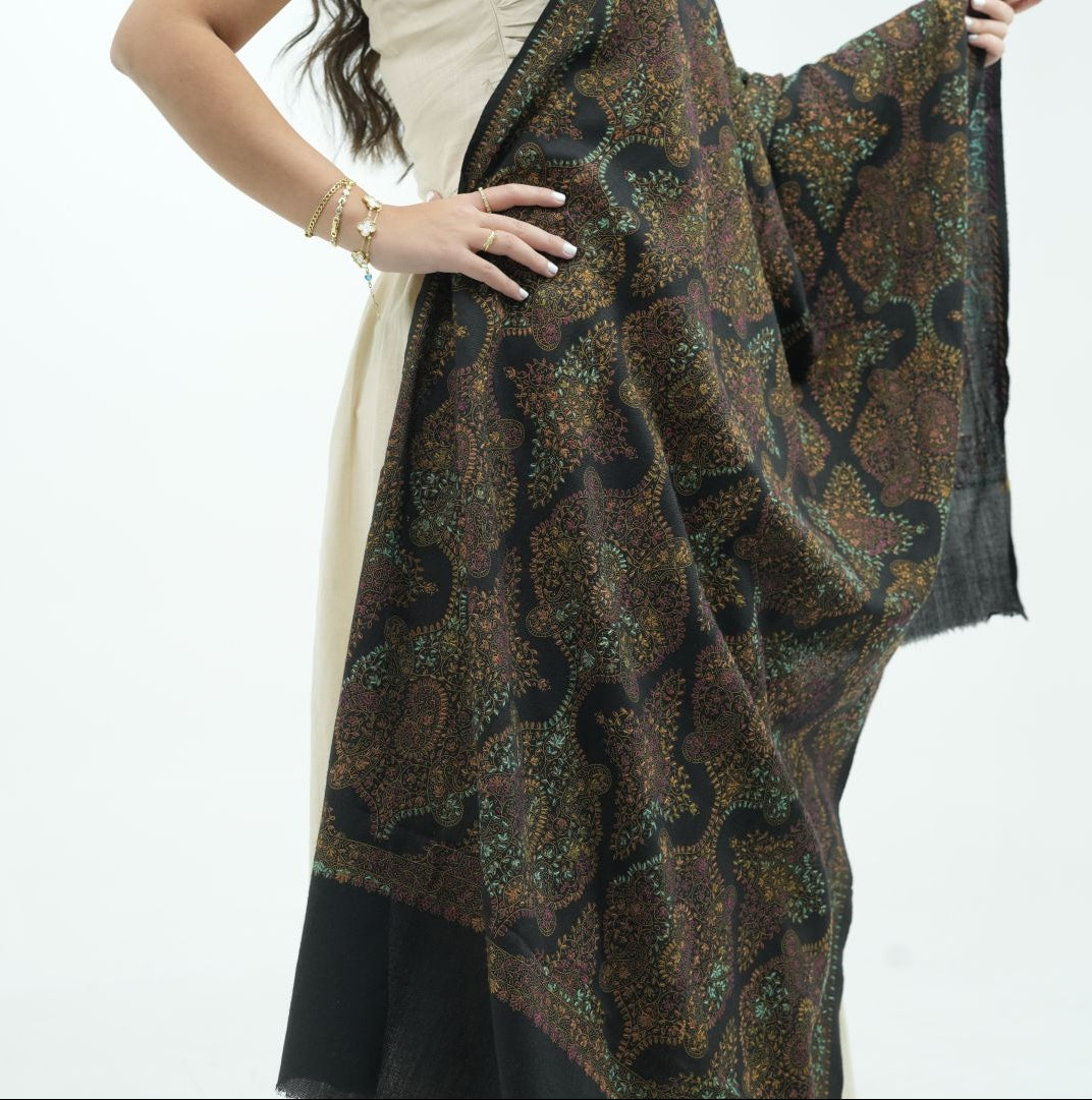 A woman models a dark brown and black shawl with an intricate paisley pattern, draped over her shoulders and held out to display its pattern and sheer edges against a plain white background.