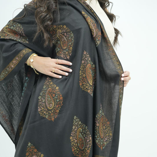 A woman wearing a black shawl with intricate brown, gold, and green paisley patterns, shown from the side against a plain white background.