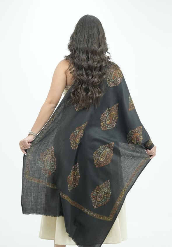 A woman stands with her back to the camera in a white studio, holding up a dark charcoal gray dupatta with gold and brown paisley patterns. She is wearing a cream-colored dress and black heels.