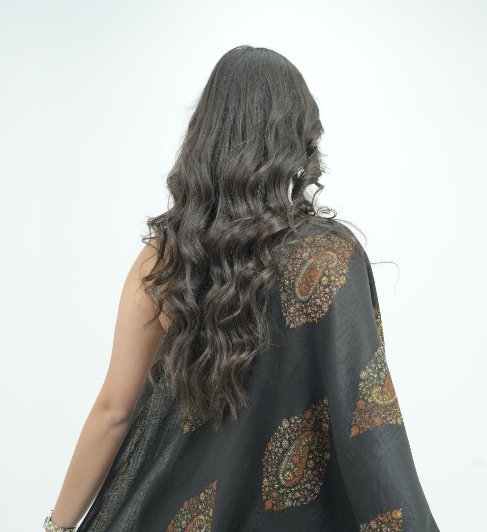 A woman with long, wavy dark brown hair is shown from behind, wearing a black shawl with an intricate gold, brown, and orange paisley pattern against a plain white background.